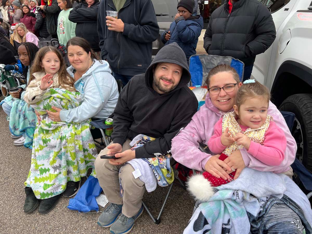 People seated outdoors in folding chairs, smiling and holding young children, with a crowd behind them.