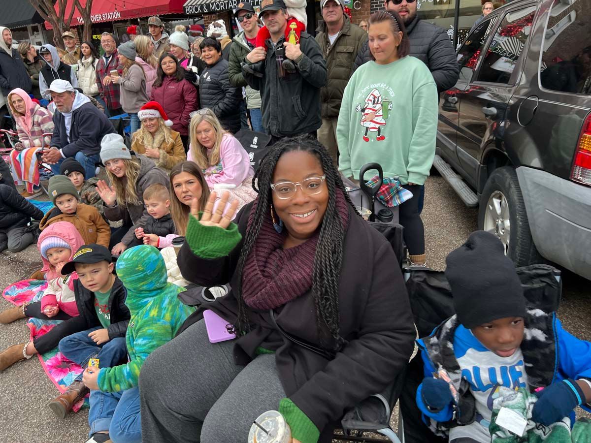 Crowd seated along a curb at a parade, with a smiling woman waving in the foreground