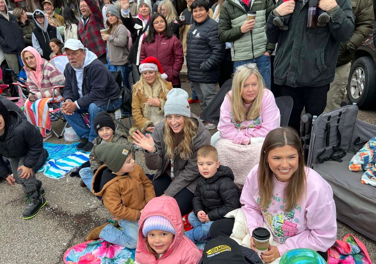 Crowd seated outdoors on a sidewalk, bundled in coats and hats, watching an event nearby