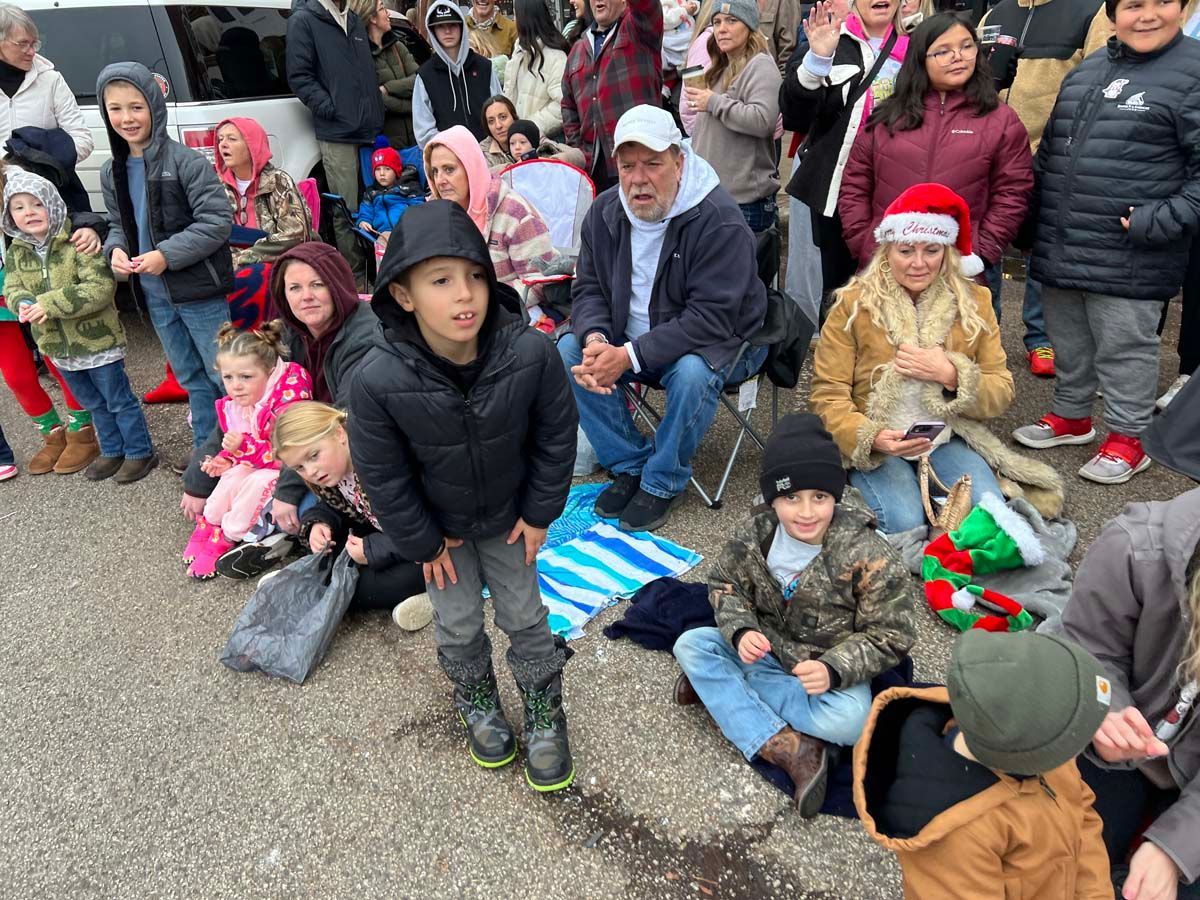 Children and adults sitting together outdoors, many in winter clothes and festive hats, watching a street event