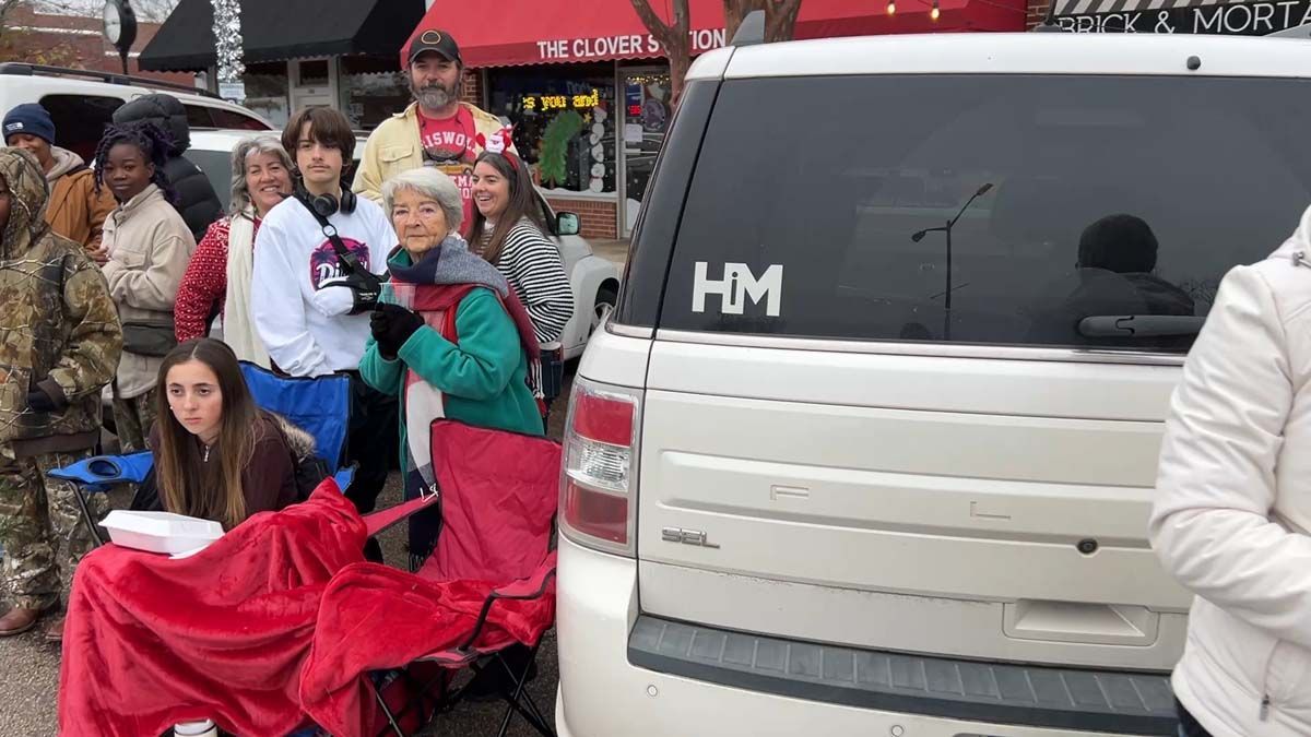 People waiting beside a white van in a crowded outdoor scene, one seated in a red blanket.