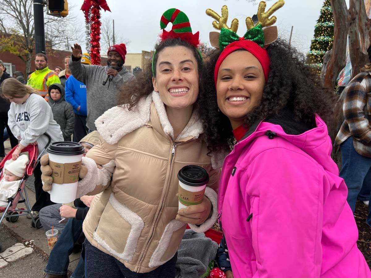 Two women in festive headbands smiling and holding coffee cups outdoors at a holiday event