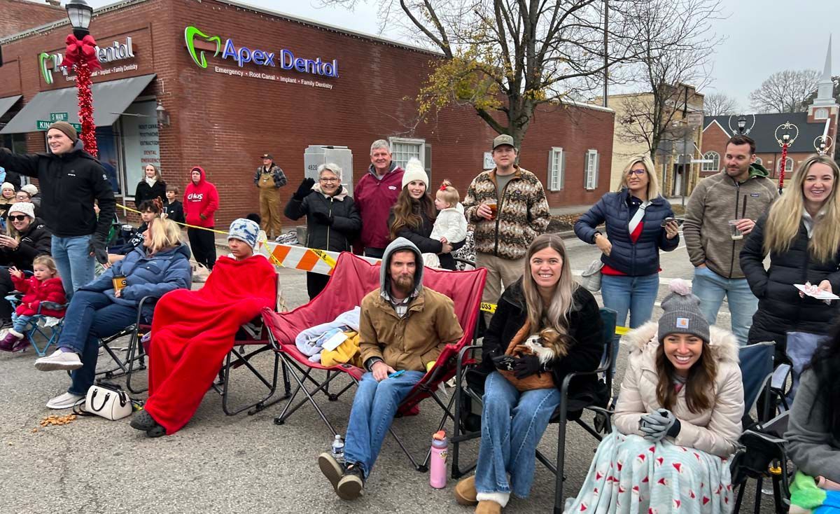 Crowd gathered outdoors by a brick building, many seated in folding chairs and smiling at the camera.