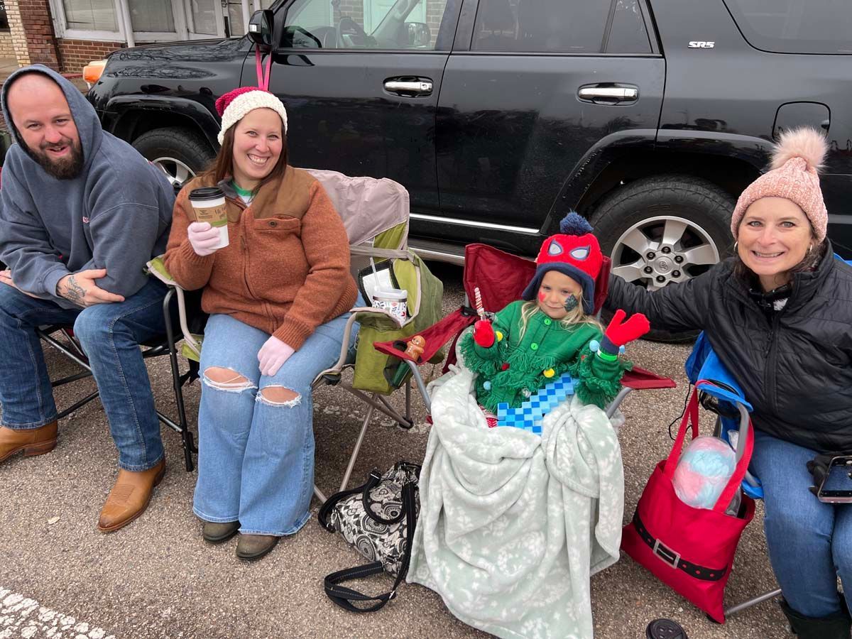 Four people seated outdoors beside a black SUV, with a child in a red hat and holiday decorations in front.