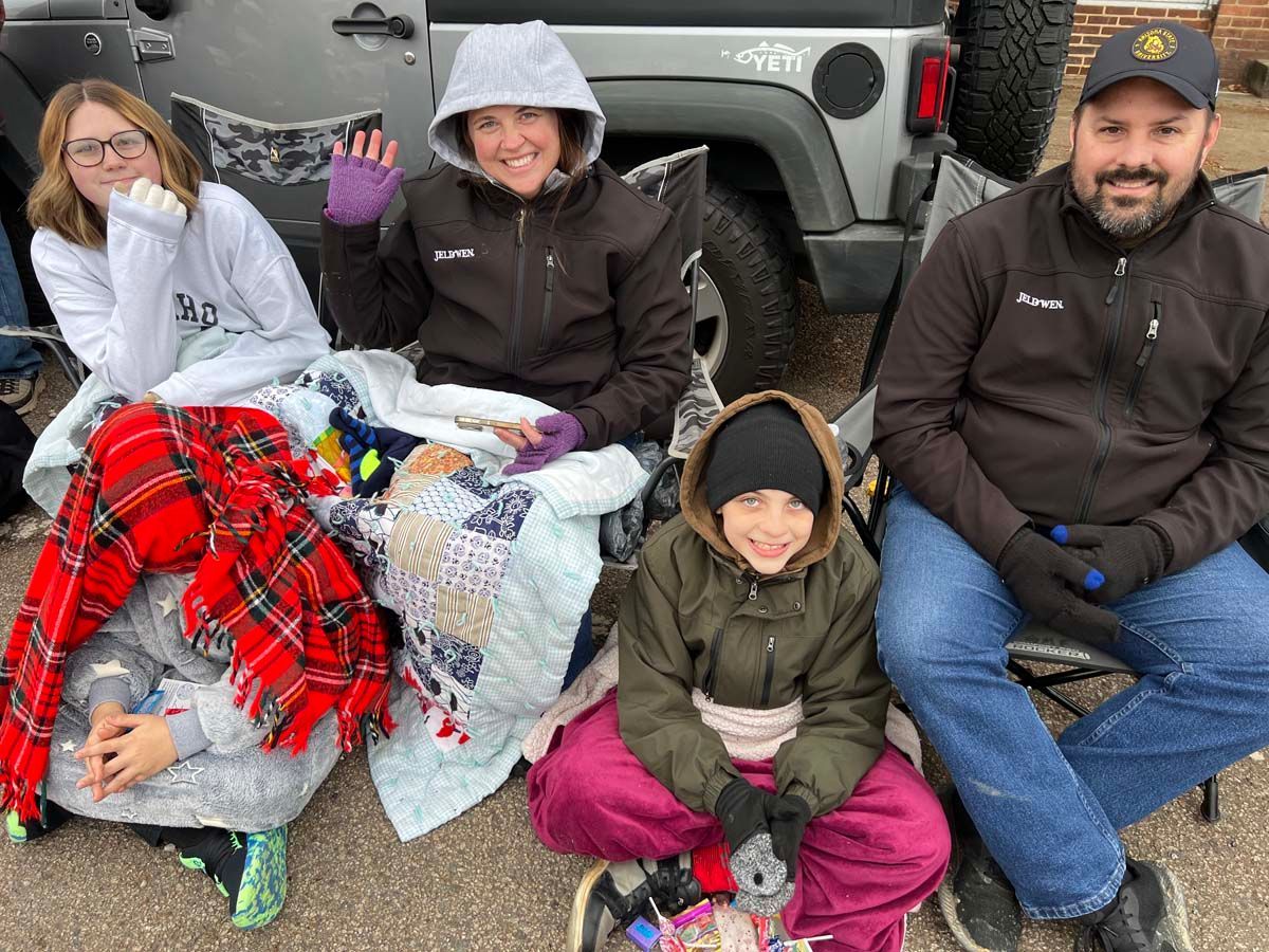 Five people sitting on the ground in winter clothes, smiling in front of a gray Jeep.