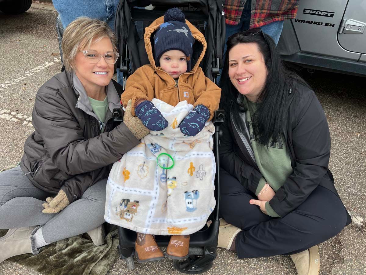 Two women crouch beside a bundled toddler in winter clothes outdoors by a parked car.
