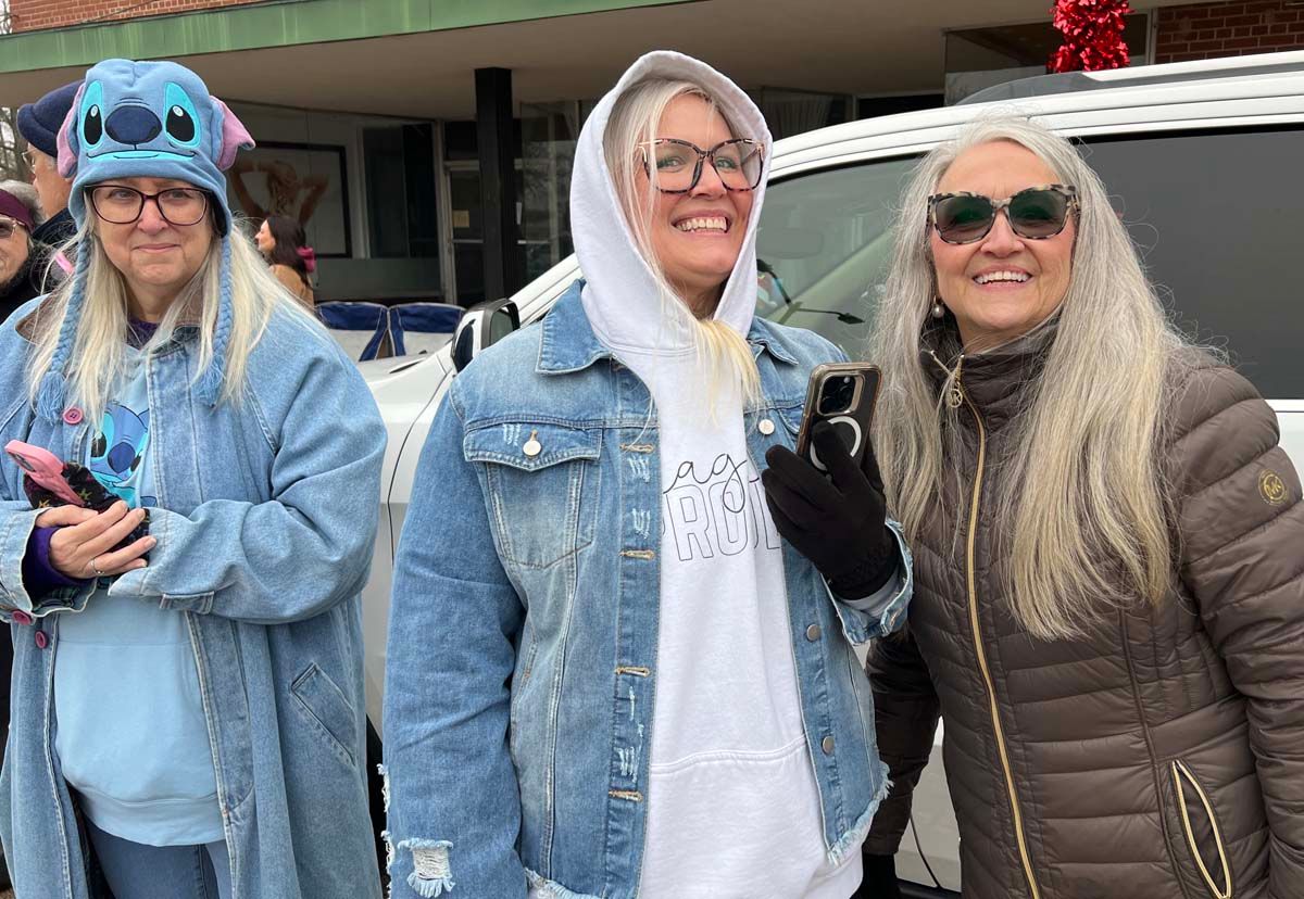 Three people in winter coats and hoodies smiling outdoors in front of a vehicle.