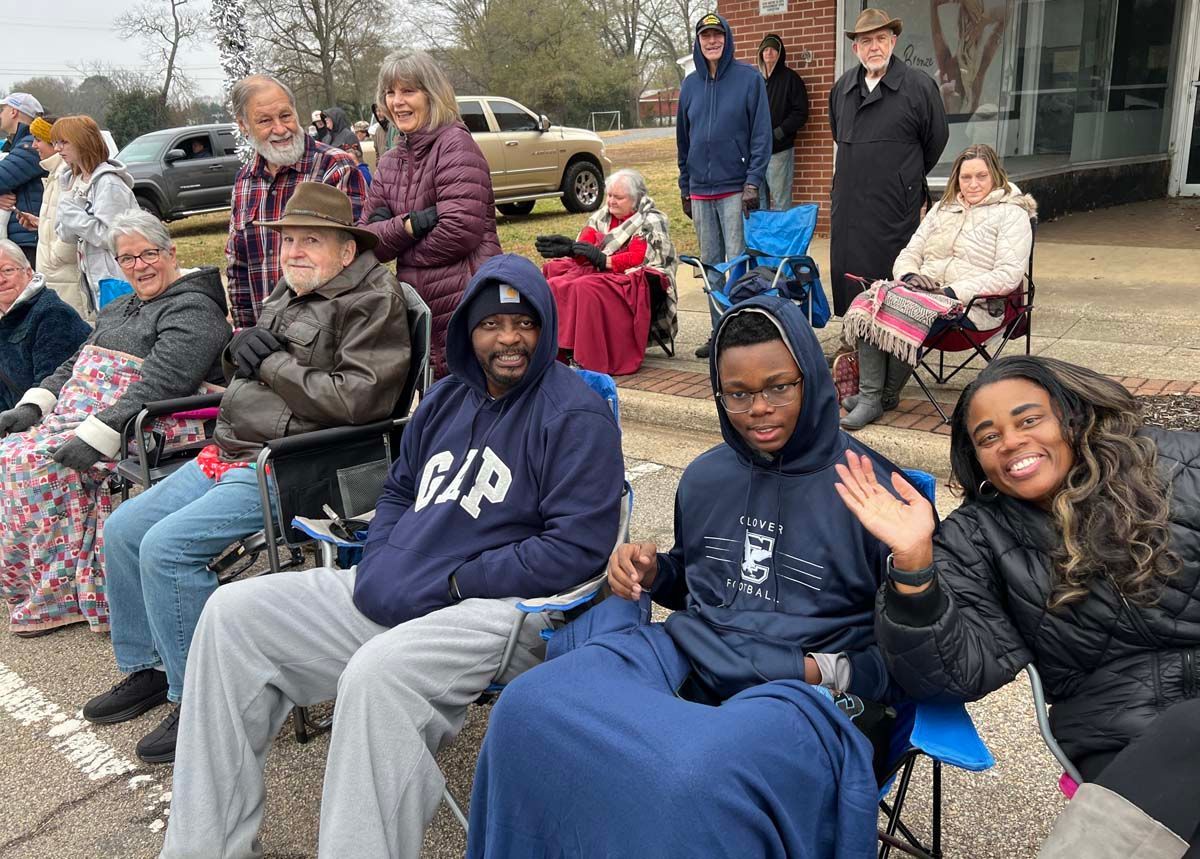 People seated outdoors in folding chairs, some smiling and waving in a neighborhood gathering