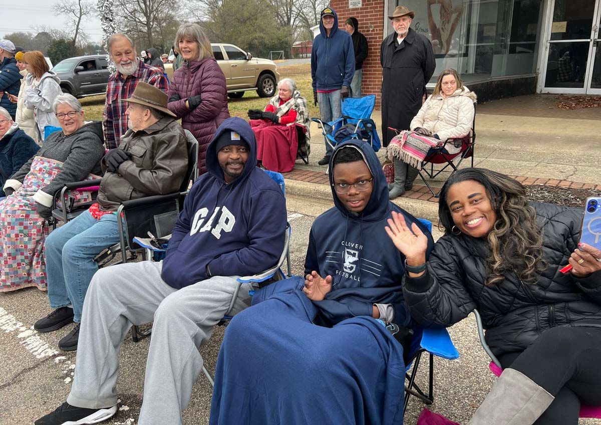 People seated outdoors on benches, smiling and waving near a building.