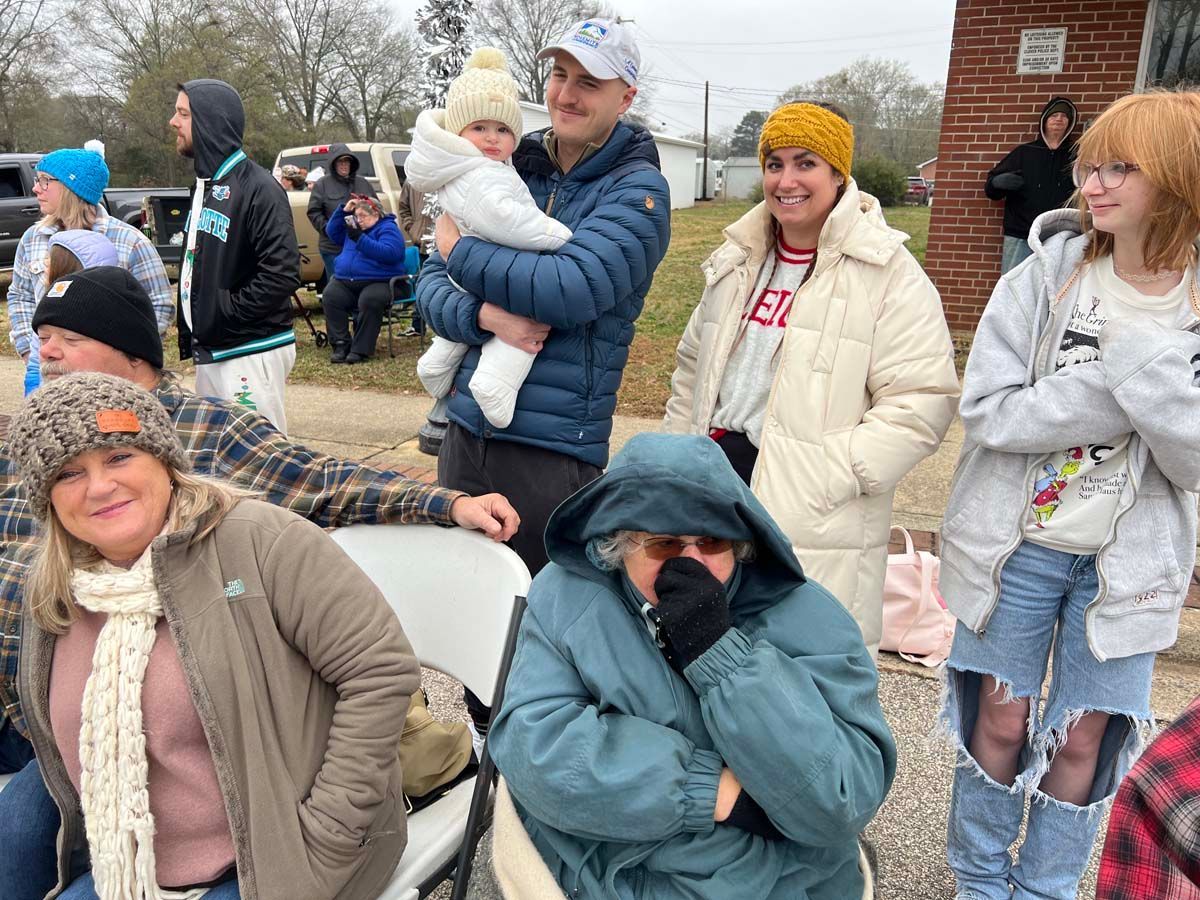 People in winter coats gathered outdoors, some seated and some standing near a brick building.