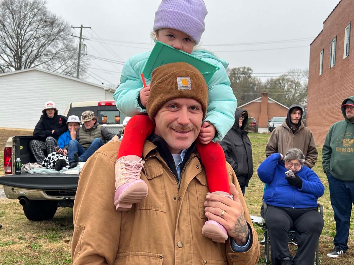 Man carrying a child on his shoulders at an outdoor winter gathering, with people and vehicles in the background