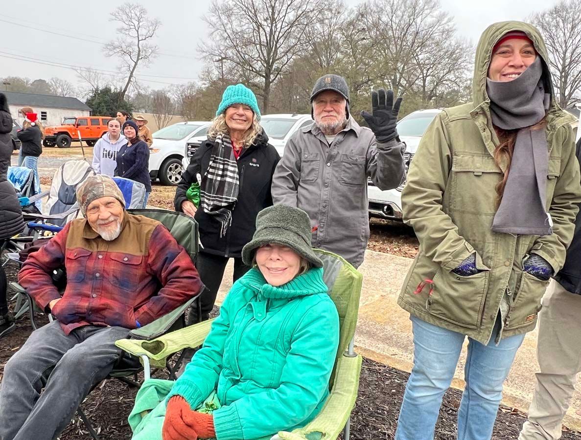Small group in winter coats gathered outdoors, smiling and seated near a parked car on a chilly day.