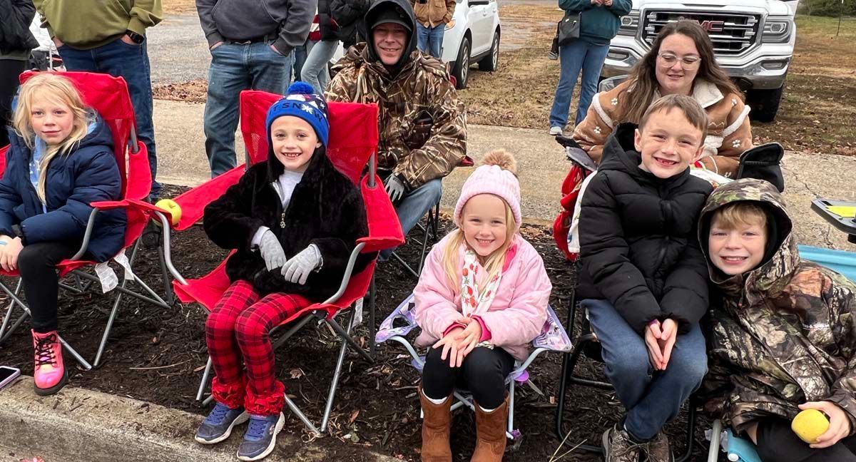 Children sitting in folding chairs outdoors, bundled in coats and smiling in a chilly setting.
