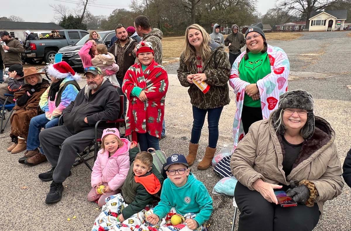 A group of people seated and standing outdoors in winter clothing, smiling in a parking lot.