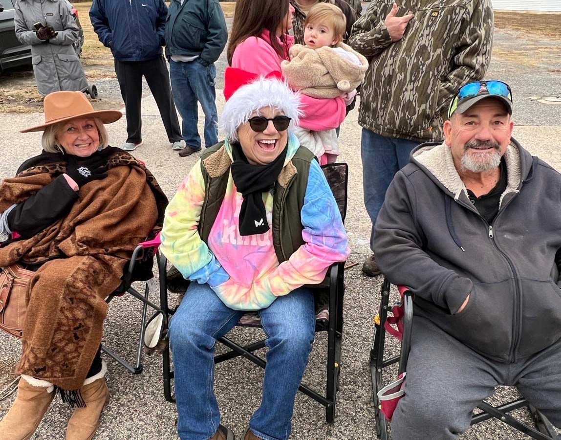 Three people in folding chairs outdoors, smiling at a gathering; one wears a tie-dye shirt and Santa hat.