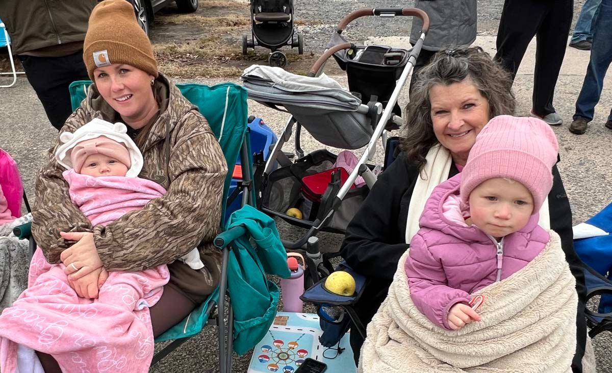 Two adults with bundled toddlers in strollers at an outdoor event, wearing winter hats and coats.