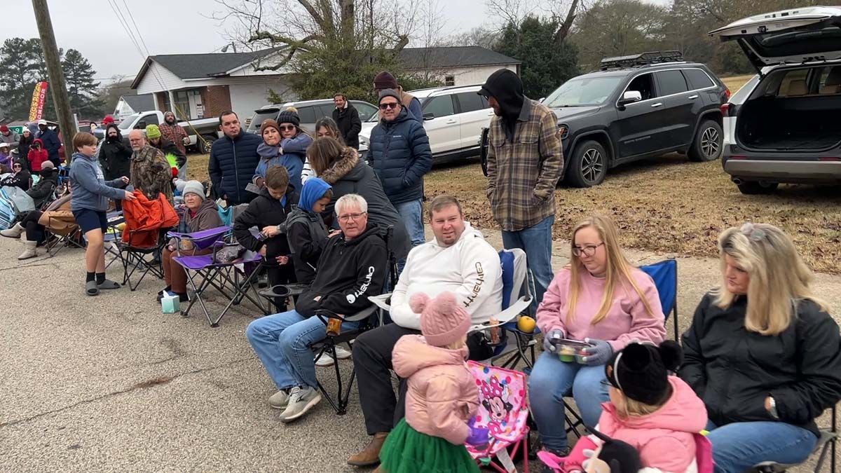 People seated in folding chairs along a street, watching a parade on a cloudy day.