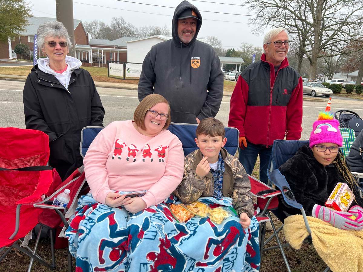 Five people posing outdoors with two children seated in red folding chairs and blankets on a chilly day