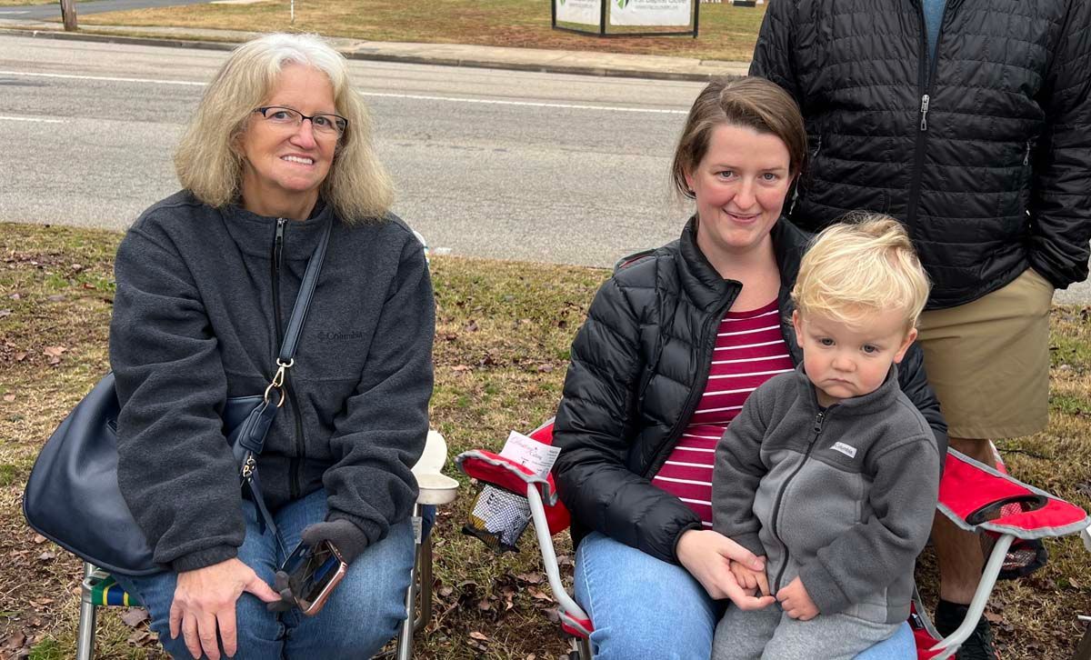 Two people seated outdoors in folding chairs, one holding a toddler, with a sidewalk and trees behind them.