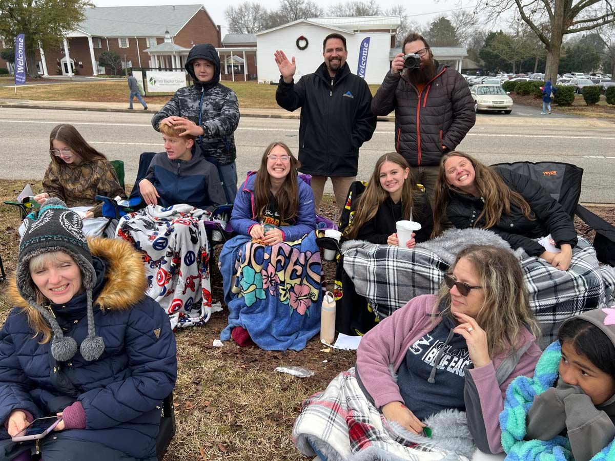 Group of people sitting outdoors on blankets in winter coats, posing and smiling near a road.