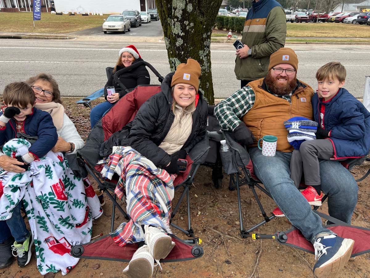 People seated outdoors in camping chairs and blankets, smiling on a cold day near parked cars.