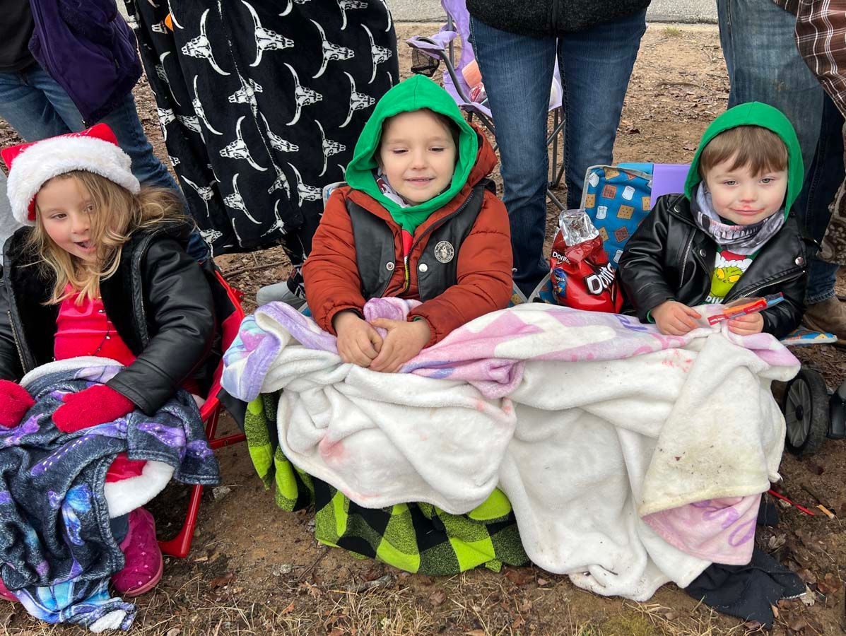 Three children bundled in blankets and hats, sitting outdoors on a bench with wrapped gifts nearby