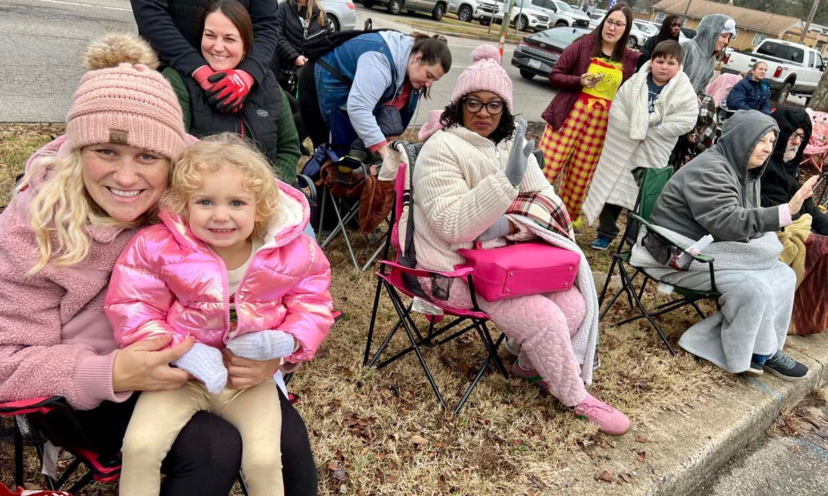 Crowd seated outdoors in folding chairs, bundled in winter clothing, some smiling at a parade or event