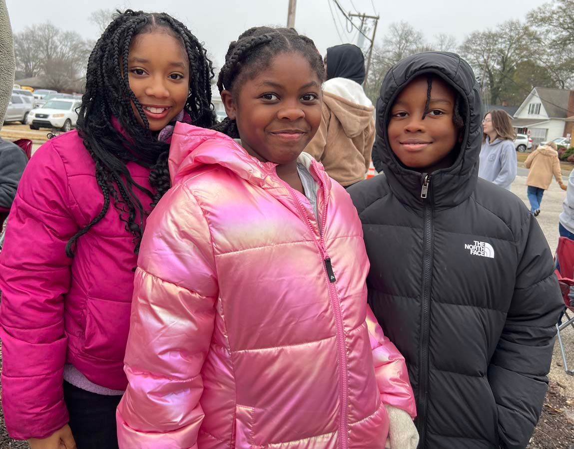 Three people in puffy winter jackets smile outdoors on an overcast day.