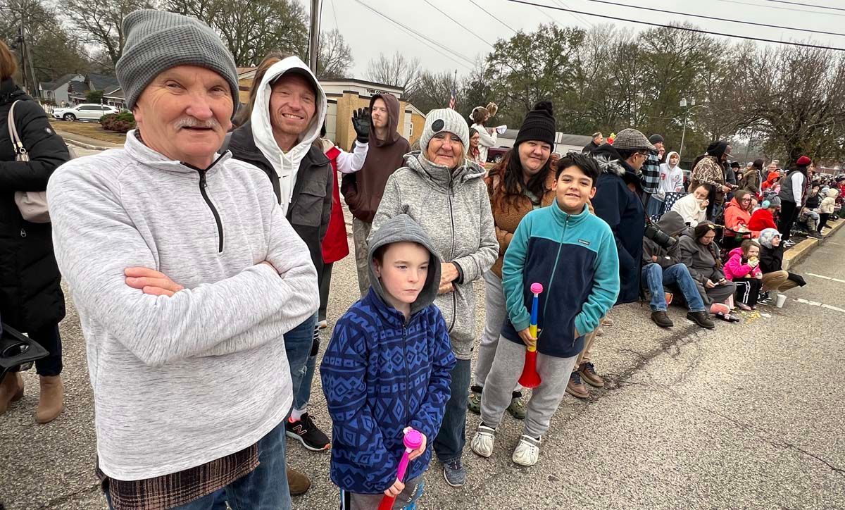 People lined up along a roadside on a cloudy day, some wearing winter hats and holding colorful ribbons.
