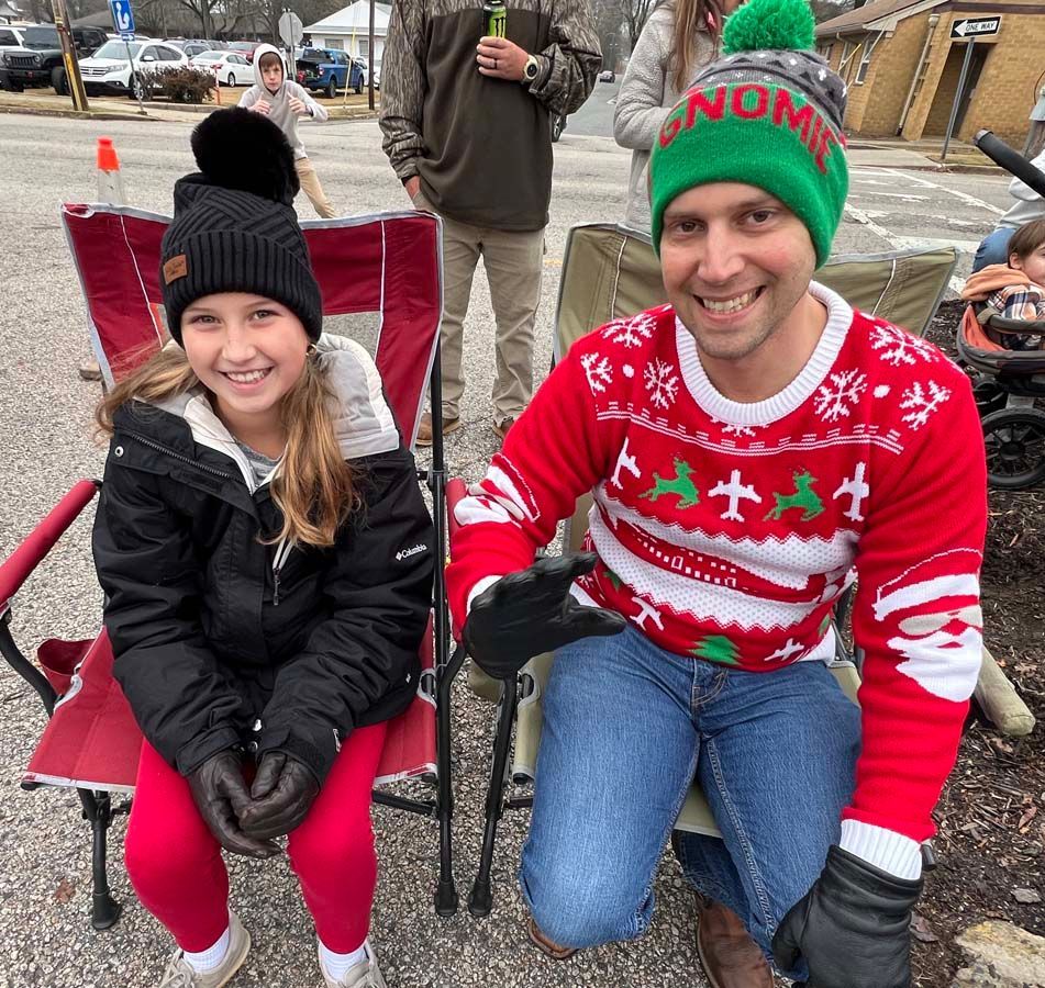 Two people smiling outdoors in winter clothes, seated in folding chairs, one wearing a red holiday sweater and elf hat.
