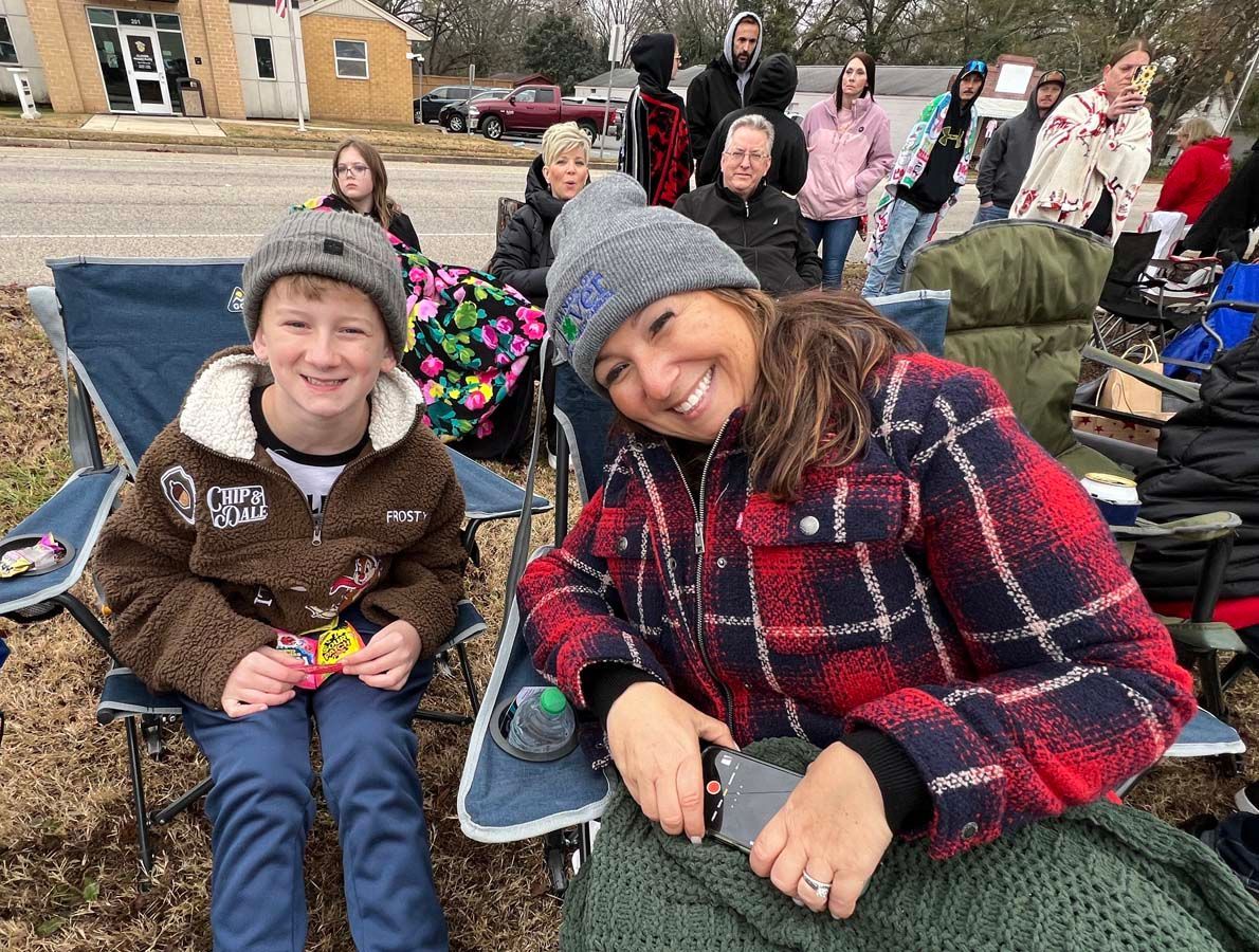 Two smiling children in folding chairs at an outdoor gathering, with people seated behind them.