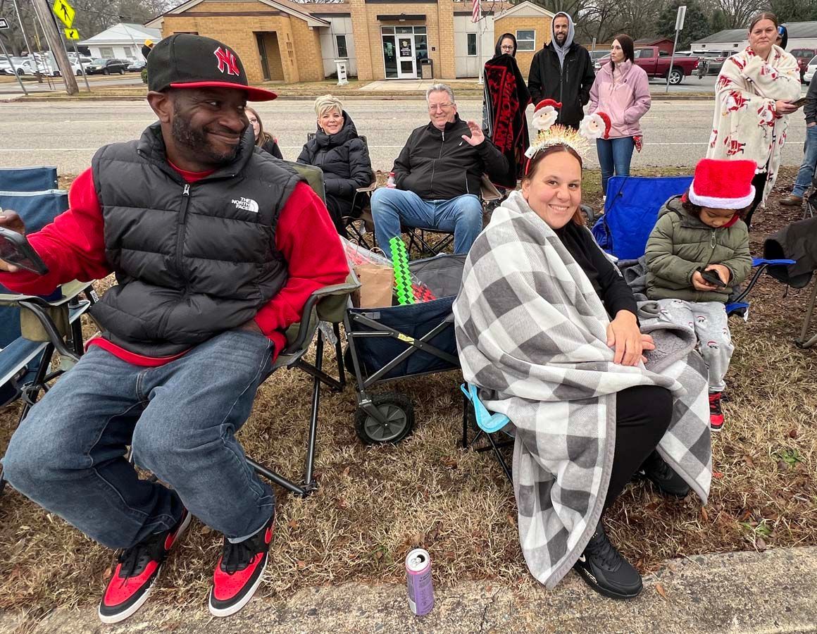 People seated outdoors by a curb, wrapped in blankets, with folding chairs and a small crowd behind them.