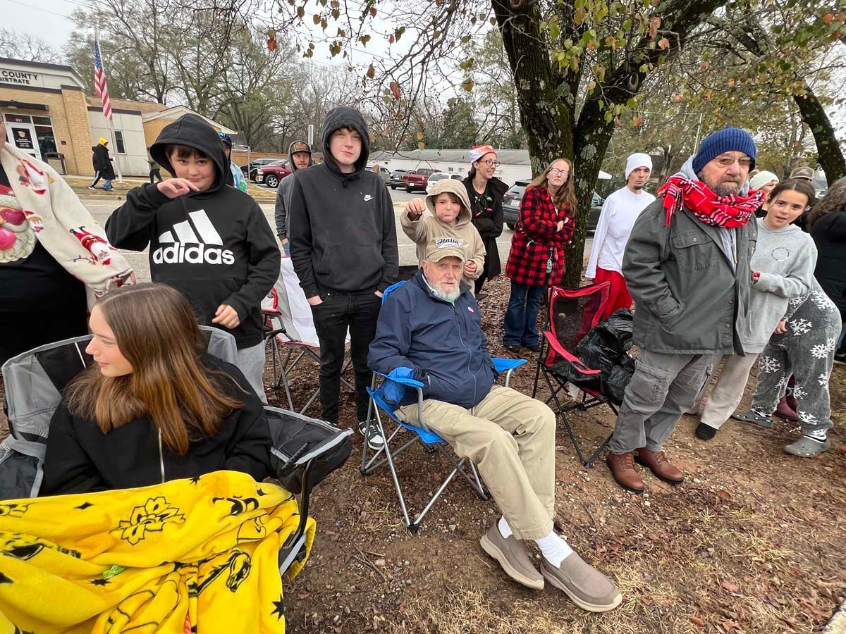 Group of people seated outdoors on a chilly day, bundled in coats and hats near a tree and parked cars.