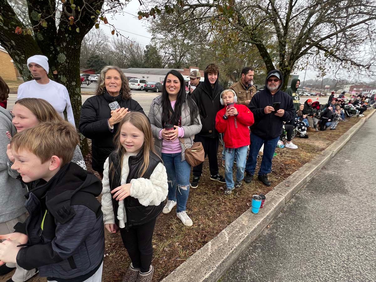 People lined along a curb outdoors, smiling and watching a parade route on a cloudy day.