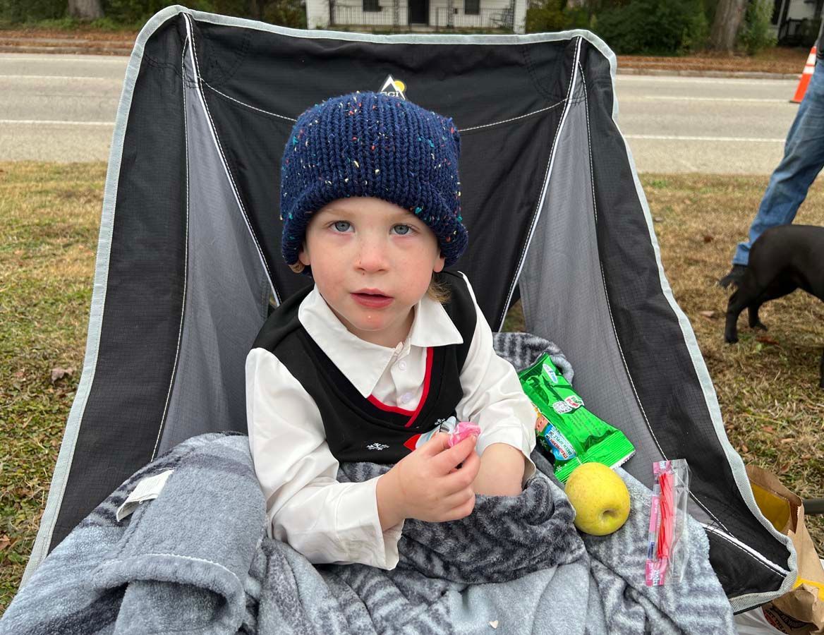 Child in a navy knit hat sits in a stroller with a gray blanket and toy, outdoors on grass.