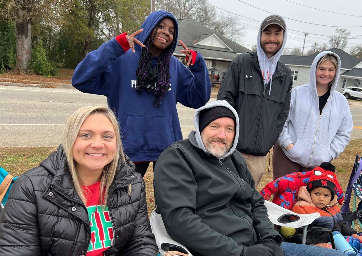 Six people gathered outdoors on a cloudy day, smiling and posing near a street with trees and houses.