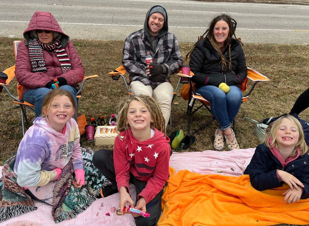Seven people in camping chairs and blankets, smiling outdoors in a grassy field.