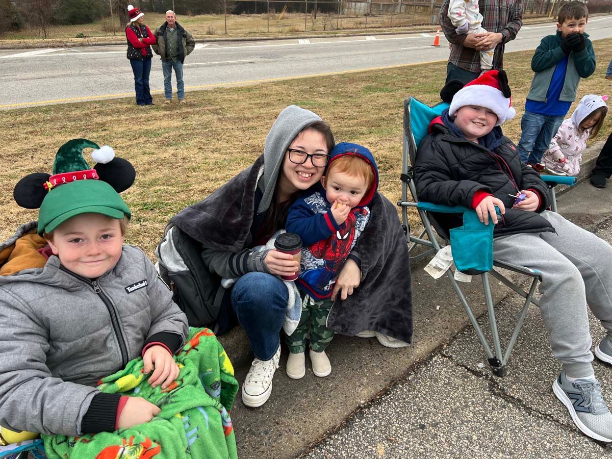 Children in holiday hats and coats sit together outdoors, smiling on a curb with a stroller nearby.