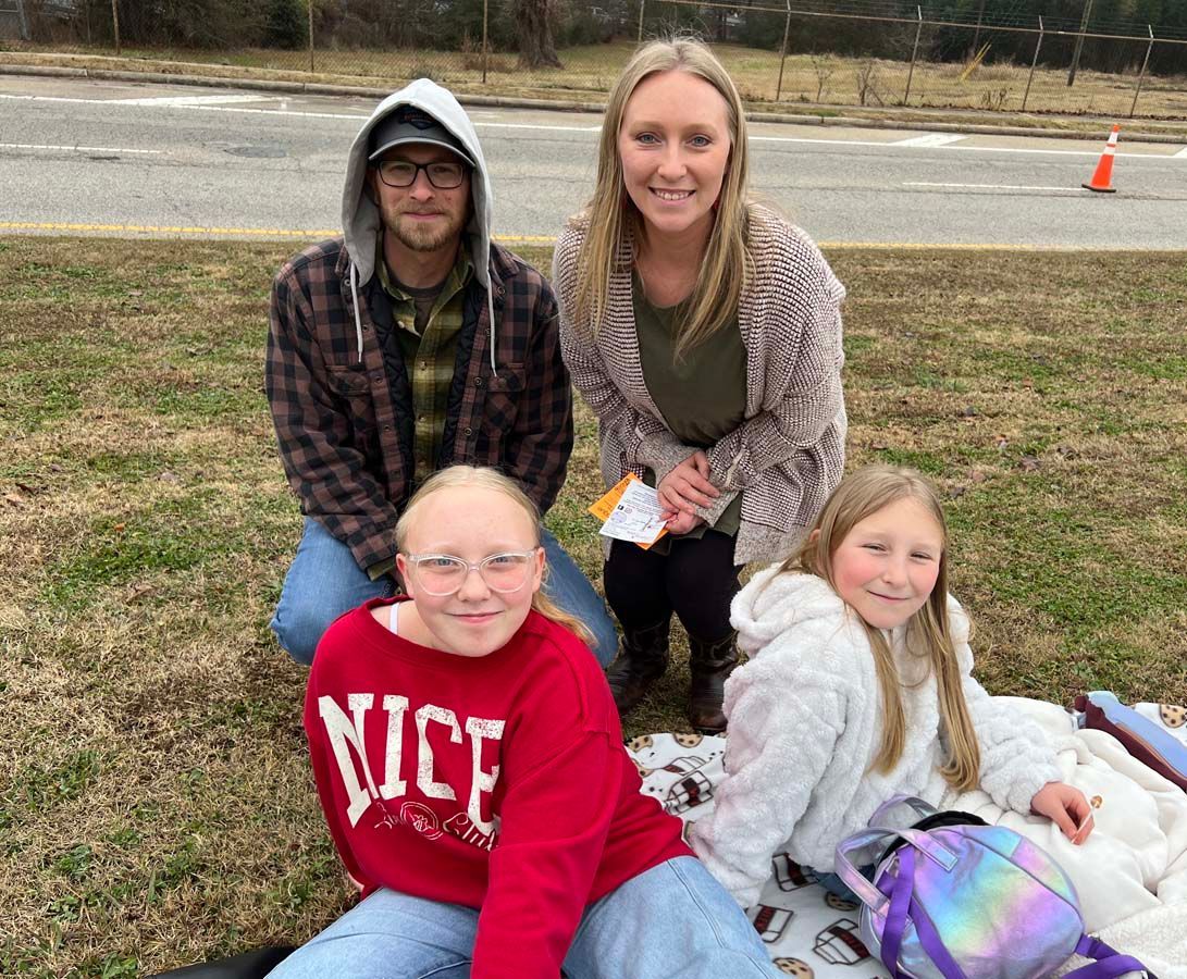 Four people posing outdoors on grass near a road, two standing behind two seated, smiling.