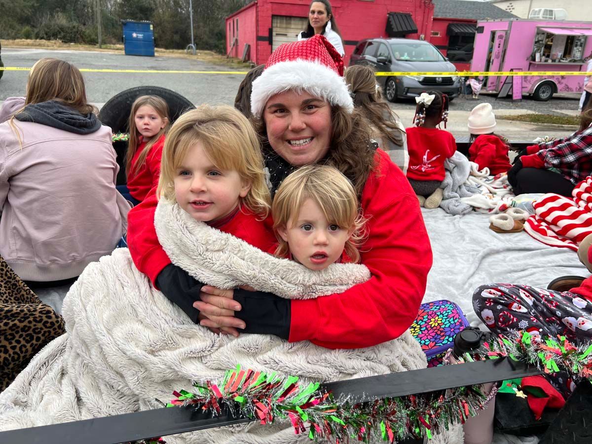 Woman in Santa hat hugging two children wrapped in blankets at a festive outdoor holiday event