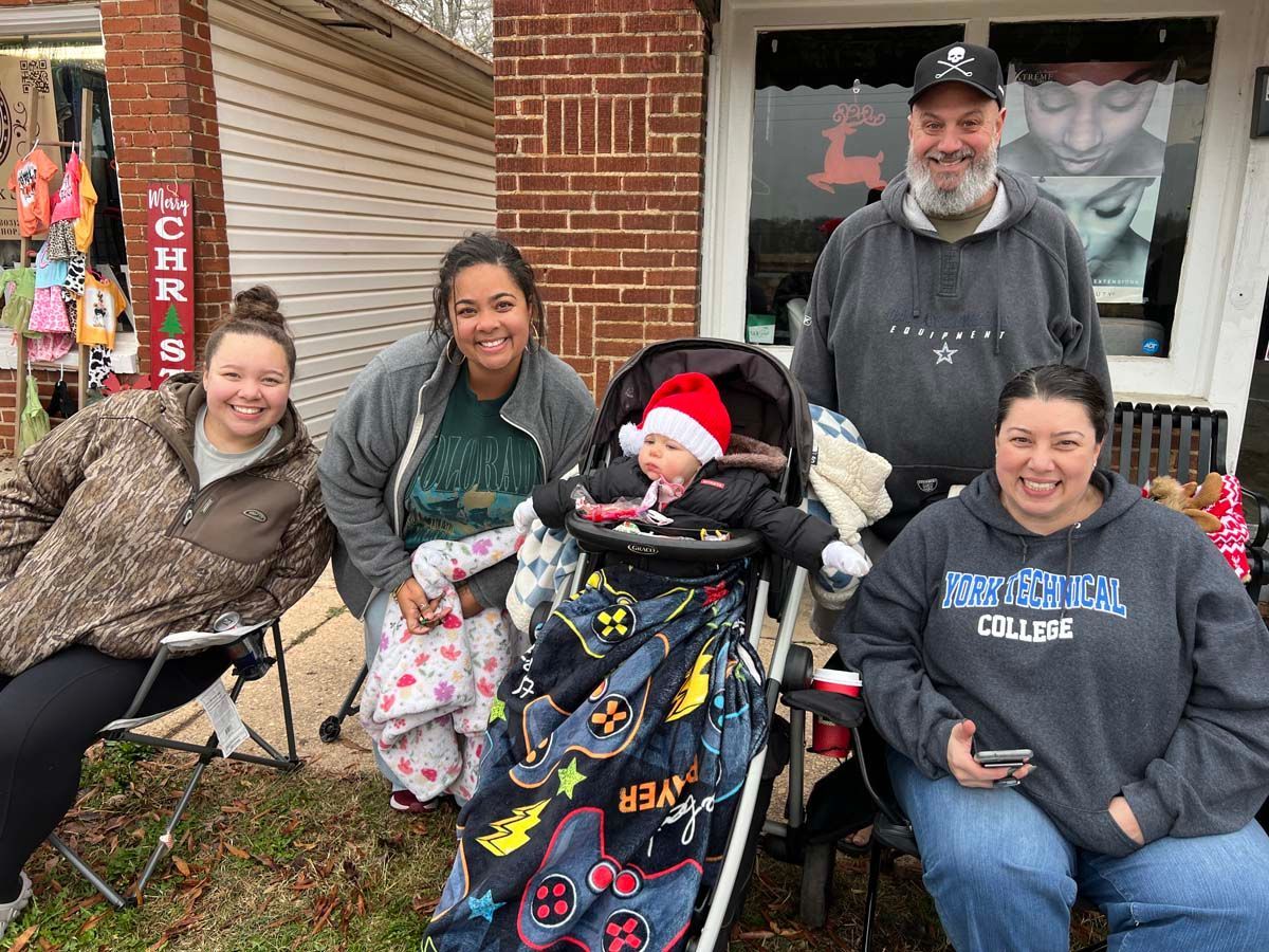 Four people pose outdoors with a child in a stroller, in front of a brick building.