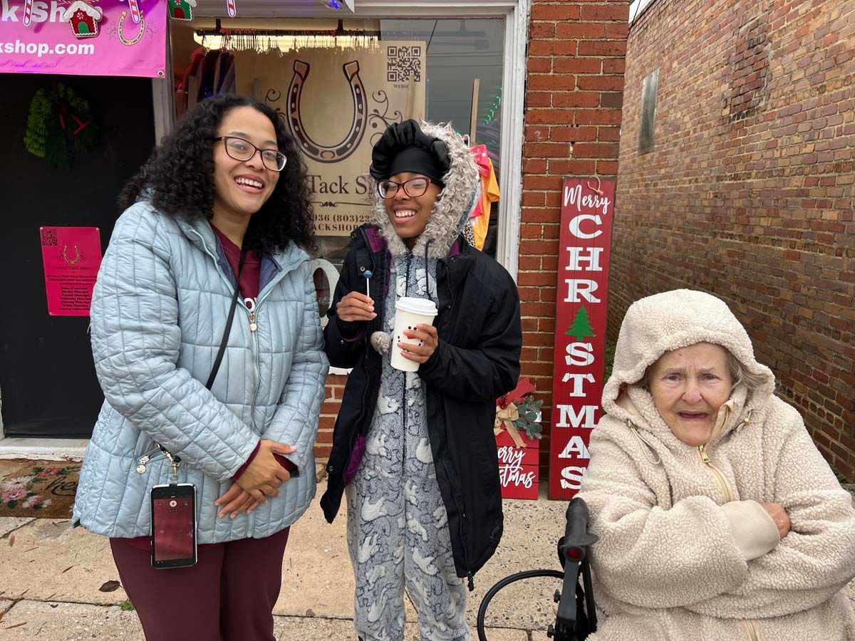 Three people smiling outside a shop, one holding a cup, with a Christmas sign and decorations nearby