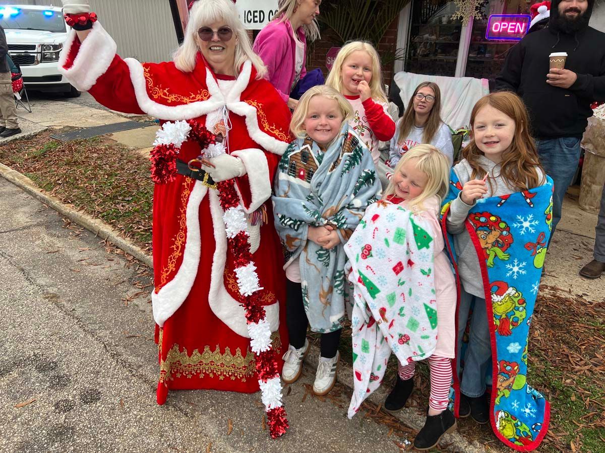 Woman in red Santa-style dress posing with four children outside a store, smiling by an open sign.
