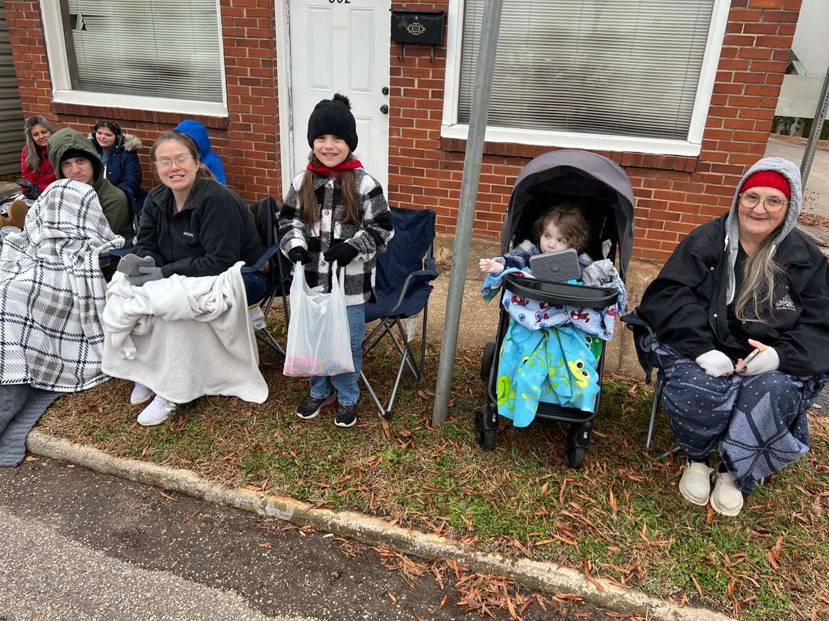 Five people seated outdoors by a brick building, wearing winter clothes and sitting with blankets and a stroller.