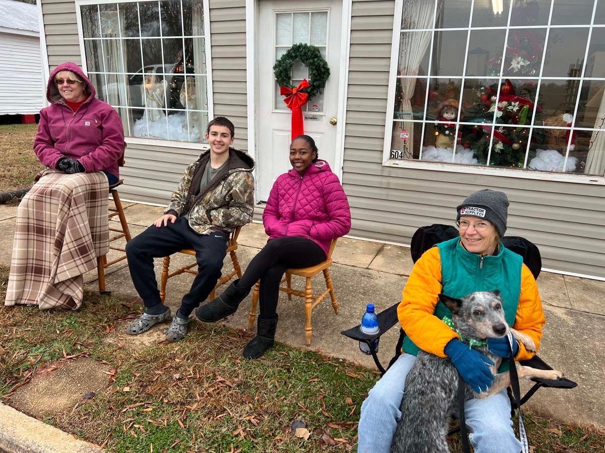 Four people seated outdoors in front of a house, wearing winter clothes, with a dog on one lap.
