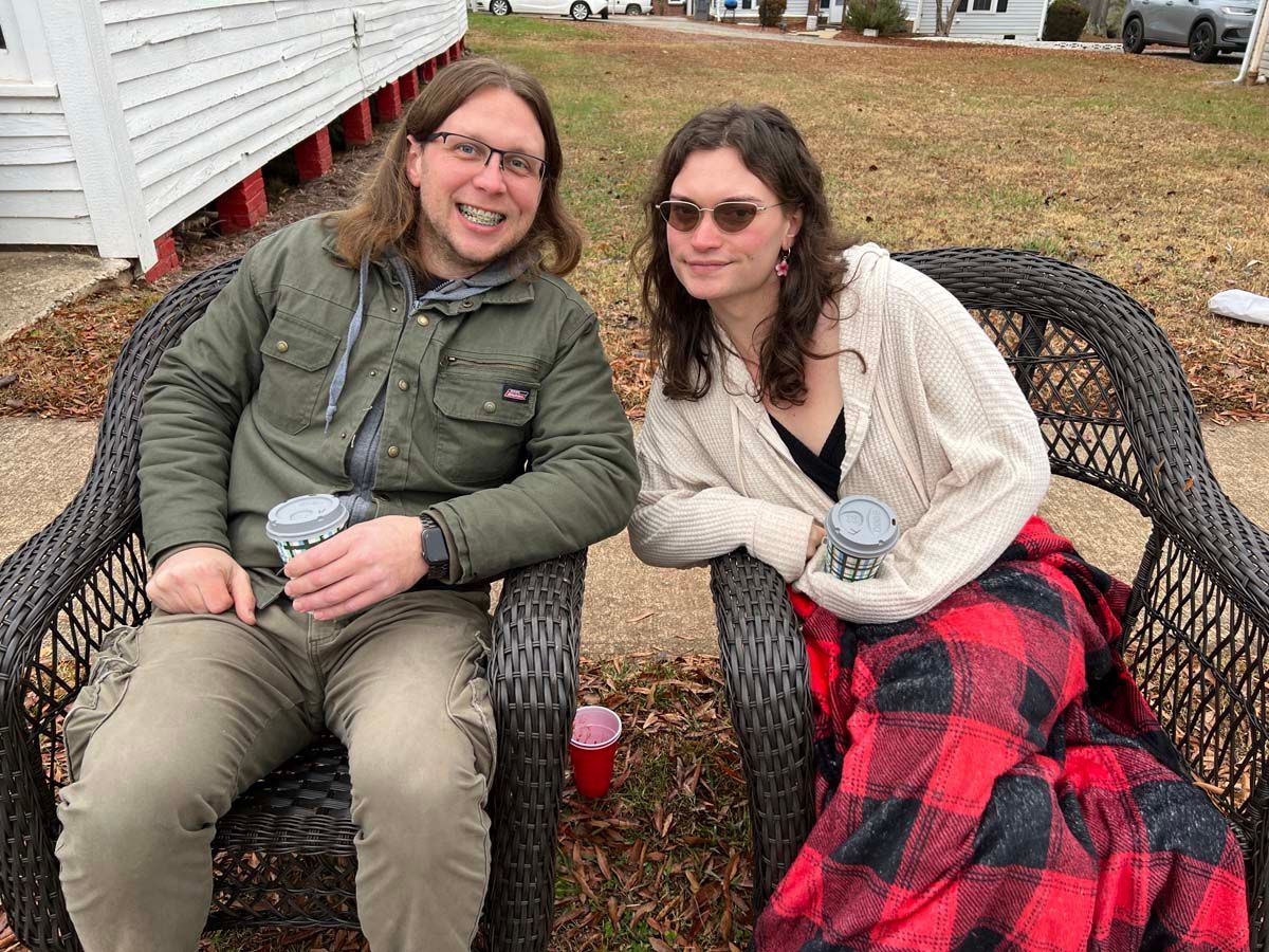 Two people sit outdoors in wicker chairs, smiling and holding drinks on a grassy lawn.