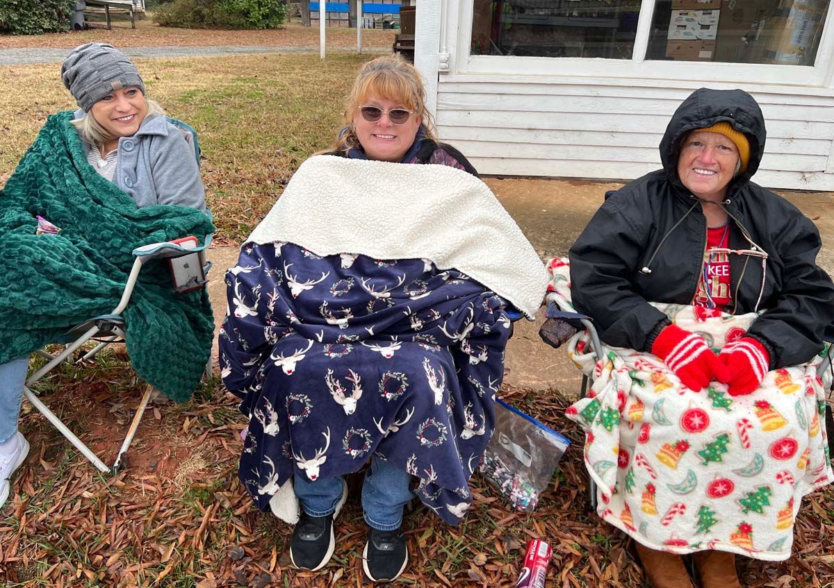 Three people seated outdoors on folding chairs, wrapped in blankets, smiling in front of a house