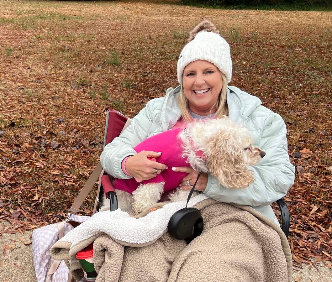 Smiling woman in white hat holding a small dog in a pink sweater on a blanket outdoors in autumn.