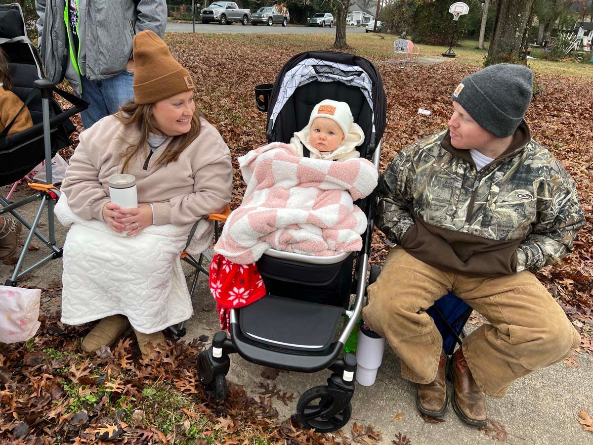 Three people sitting outdoors on a leaf-covered sidewalk, with a baby in a stroller wrapped in a pink blanket.