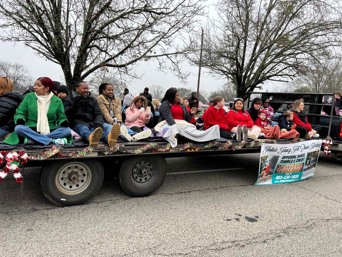 People seated on a trailer float during a parade, wearing colorful clothes and waving.